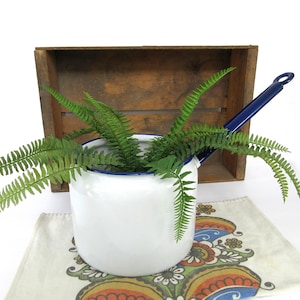 May include: A white enamel pot with a dark blue rim and a long matching handle. The pot features a cylindrical body and contains a green fern plant with multiple fronds extending outward from the center of the vessel, creating a natural display.