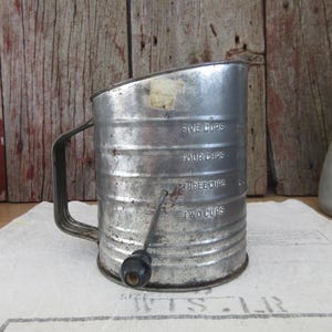 May include: Vintage silver metal measuring cup with a black handle and a rotating sifter. The cup has markings for two, three, four, and five cups.