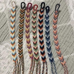 Könnte beinhalten: Eine Sammlung von sieben handgefertigten Freundschaftsarmbändern. Jedes Armband hat ein Zickzackmuster in verschiedenen Farben, darunter Blau, Orange, Braun, Grün und Rosa. Die Armbänder haben Schlaufenenden und geflochtene Quasten.