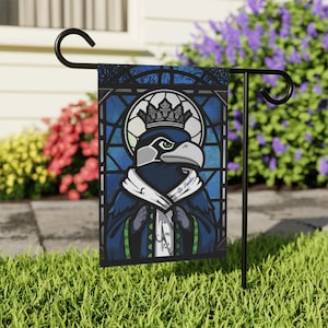 May include: A rectangular garden flag featuring a stained glass style illustration of a bird wearing a crown and a cape. The bird has dark blue feathers, a grey beak, and green accents. The text "Go Hawks" and "We are 12" is visible on the cape.