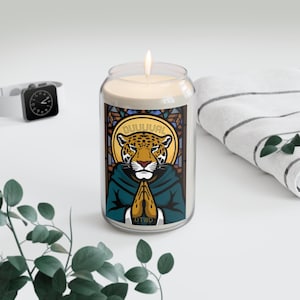 May include: A lit candle in a glass jar with a label featuring a jaguar illustration with the text "DUUUVAL" and "DTWD". The jaguar is depicted with folded hands and a halo. The candle is on a white surface with a watch and a towel.