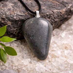 Peut inclure: Un collier pendentif en forme de goutte, gris foncé, avec une bélière argentée. Le pendentif poli est suspendu à un cordon noir. Le collier repose sur un lit de cristaux blancs et une roche gris foncé.