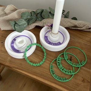 Puede incluir: Dos portavelas circulares blancos con textura acanalada. Uno sostiene una vela blanca. Se incluyen anillos morados con el texto en alemán "HERZMENSCH" y "DU BIST WUNDERVOLL". Tres anillos verdes presentan motivos de hojas y el texto "SCHÖN DASS ES DICH GIBT".