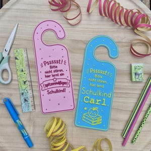 May include: Two door hangers in pink and blue with German text, accompanied by school supplies including scissors, a ruler, a blue pen, a dinosaur eraser, two pencils, and decorative curled ribbons arranged on a round wooden tray.