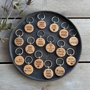 Puede incluir: Una colección de llaveros redondos de madera con anillas metálicas, cada uno grabado con texto en alemán y pequeños símbolos. Los llaveros están dispuestos sobre una bandeja circular de color gris oscuro. El texto incluye Bester Trainer y Lieblings Trainerin.