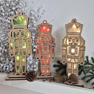 May include: Three wooden nutcracker figurines with internal lights in green, red, and white. Each nutcracker stands on a wooden base and has a crown. The background includes a white wreath and pinecones, suggesting a holiday theme.