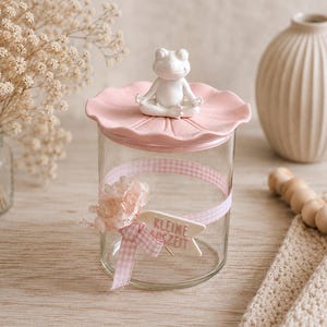 Può includere: Un barattolo in vetro trasparente con coperchio rosa sormontato da una statuetta di rana bianca in posizione di meditazione. Il barattolo è decorato con un nastro a quadretti rosa e bianco, fiori secchi e un'etichetta con la scritta KLEINE AUSZEIT.