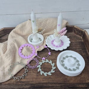 May include: Two white taper candles with small heart decals, each placed in a white circular candle holder. One holder has a scalloped edge. Included are five flat, circular decorative rings in shades of sage green and light purple, featuring heart-shaped cutouts.