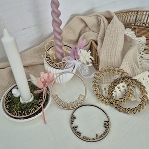 Puede incluir: Una colección de elementos decorativos, que incluyen velas y adornos de madera. Una vela alta y retorcida de color lavanda se encuentra en una maceta blanca con flores secas. También hay dos velas blancas, junto con anillos de madera con texto y diseños de hojas.