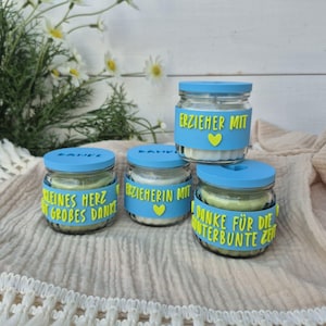 Puede incluir: Cuatro pequeños tarros de cristal con tapas y etiquetas azules. Las etiquetas tienen texto en alemán y un corazón amarillo. Los tarros están sobre una tela crema, con margaritas en el fondo. Tarros de regalo.