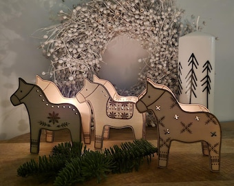 May include: Three wooden horse candle holders with decorative cutouts, each with a unique design. A white wreath and a candle with black tree silhouettes are in the background. A sprig of evergreen is in front of the horses.