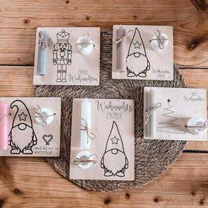 Puede incluir: Adornos navideños de madera con velas, con un cascanueces, gnomos y un diseño de árbol de Navidad. Cada adorno incluye una vela, un elemento decorativo y texto en alemán. Los adornos están dispuestos sobre una superficie de madera.