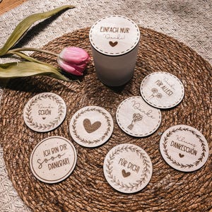 Puede incluir: Un juego de posavasos de madera y un pequeño recipiente. Los posavasos tienen varias frases y diseños de corazón. La tapa del recipiente dice "EINFACH NUR Danke!" con un corazón. También hay un tulipán rosa y vegetación.