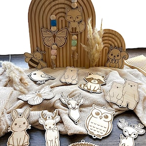 Op de afbeelding: Houten mobiel voor de kinderkamer met uitgesneden bosdieren, waaronder een vos, een vlinder en een beer. De mobiel heeft een regenboogvormige houten basis en hangende kralen. Extra houten dierenuitsparingen zijn rond de basis geplaatst.