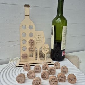 May include: A wooden wine bottle-shaped display stand with several round holes and a wine glass cutout. It holds small wooden wine charms with phrases like "Cheers" and "Wine Not?" A green wine bottle sits next to the stand. The theme is wine and celebrations.