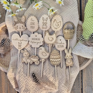 May include: Wooden Easter-themed decorations featuring bunnies, eggs, chicks, and a basket. Some decorations have text like "Love You!" and "Happy Easter." The decorations are on sticks, perfect for floral arrangements or crafts.