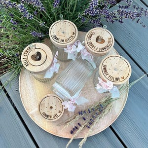 Puede incluir: Cinco vasos de cristal estriado transparente con tapas de bambú y pajitas de cristal. Las tapas presentan textos grabados como Hello Sunshine, Sonne im Herzen, Sommer & Sonnenschein, Summer Feeling y Sommerliebe, decorados con pequeñas flores de tela rosa.