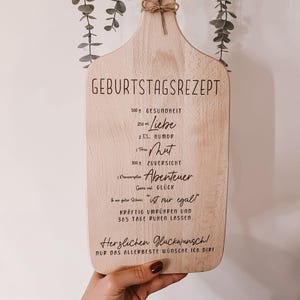 May include: Wooden cutting board with the text "GEBURTSTAGSREZEPT" and a recipe for a happy birthday. The board is decorated with dried flowers and tied with twine. The board is held by a person.