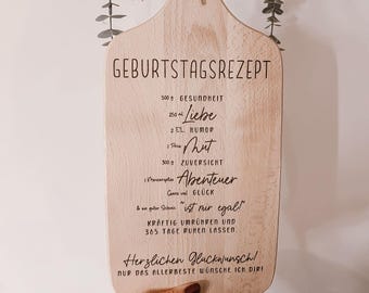 May include: Wooden cutting board with the text "GEBURTSTAGSREZEPT" and a recipe for a happy birthday. The board is decorated with dried flowers and tied with twine. The board is held by a person.
