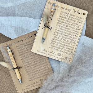 Op de afbeelding: Twee houten kaarten met de tekst "Frohes neues Jahr!" en een pen met de tekst "365 NEUE GESCHICHTEN". De kaarten hebben een lijst met woorden. Aan elke kaart is een pen bevestigd met een zwart lint. Aan elke pen is een veer bevestigd.