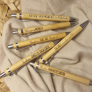 Puede incluir: Varios bolígrafos de bambú con detalles plateados, cada uno grabado con una frase alemana diferente. Los bolígrafos están colocados sobre una tela beige y una cesta de mimbre. Las frases incluyen "Te amo más que al chocolate."