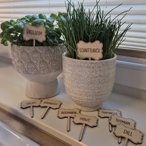 Op de afbeelding: Twee kruiden in potten, basilicum en bieslook, staan op een vensterbank. De basilicum staat in een witte, patroonpot en de bieslook in een getextureerde grijze pot. Houten plantenlabels met Duitse namen staan ervoor.