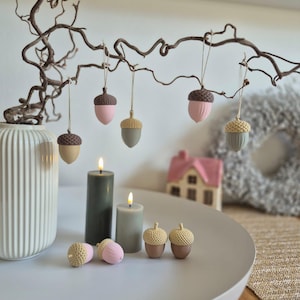 May include: A collection of decorative acorn ornaments in shades of pink, tan, and grey hanging from a twisted branch. Two pillar candles in dark green and sage grey stand on a white surface alongside four loose acorn ornaments and a white vase.