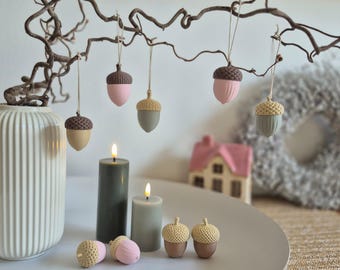 May include: A collection of decorative acorn ornaments in shades of pink, tan, and grey hanging from a twisted branch. Two pillar candles in dark and light green are placed alongside four small acorn figurines on a white circular tray.