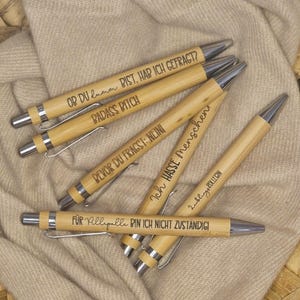 Könnte beinhalten: Eine Ansammlung von Bambusstiften mit silbernen Akzenten, die jeweils mit einem anderen Satz in Schwarz graviert sind. Die Stifte liegen auf einem beigen Stoff. Die Texte beinhalten "BADASS BITCH" und "Ich HASSE Menschen".