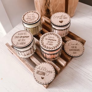 Könnte beinhalten: Vier kleine Glasbehälter mit gerippten Seiten und farbigem Wachs, jeweils mit einem runden Holzdeckel, der mit deutschem Text und Blattmotiven graviert ist. Zwei zusätzliche Holzscheiben mit Gravur liegen auf einem Holzgestell.
