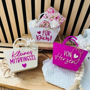 Könnte beinhalten: Drei kleine dekorative Geschenkkörbe mit Henkeln. Ein weißer Korb trägt den rosa Schriftzug "Kleines MITBRINGSEL" und ein Herz. Ein weißer Korb trägt den rosa Schriftzug "FÜR Dich!" und Blumenpapier. Ein magentafarbener Korb trägt den weißen Schriftzug "VON Herzen!" und ein Herz.