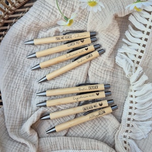 Könnte beinhalten: Eine Sammlung von Holzstiften mit schwarzen Tintenspitzen, jeweils mit deutschen Sätzen wie "Frau mit Klasse" und "Lehrer mit" graviert. Die Stifte liegen auf einem beigen Stoff, mit Gänseblümchen und einem Weidenkorb im Hintergrund.