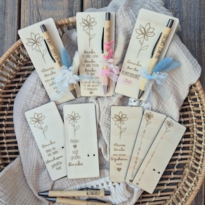 May include: Wooden pens and bookmarks with floral designs and German text. The pens and bookmarks are in a woven basket on a wooden surface. Some pens have decorative accents.