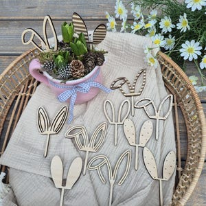 May include: A collection of wooden bunny ear decorations in various designs, alongside a pink teacup filled with greenery and pinecones. The teacup is adorned with a blue and white gingham ribbon, all arranged in a woven basket.