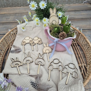 May include: A collection of wooden Easter bunny decorations. The decorations are in various shapes, including bunny heads and bottoms, and are placed on a beige cloth. A pink pot with a blue ribbon and flowers is in the background.