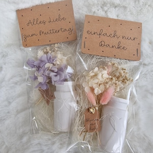 Può includere: Due piccoli bouquet di fiori secchi in confezioni di plastica trasparente, ciascuno con un vaso di ceramica bianco. Uno ha fiori viola e bianchi, l'altro fiori rosa e bianchi. Ogni bouquet ha una piccola etichetta marrone con testo tedesco.