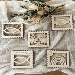 Könnte beinhalten: Fünf lasergeschnittene Holzdekorationen mit gewellten Kanten. Die Designs umfassen ein Fischsymbol, Olivenzweige, einen Regenbogen und deutschen Text. Die Dekorationen sind hellbraun und auf einem cremefarbenen Stoff angeordnet.