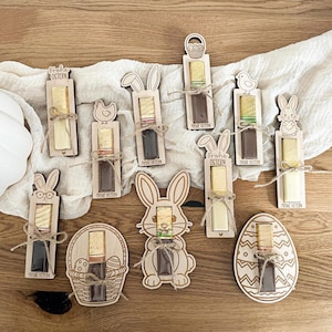 Könnte beinhalten: Osterliche Flaschenverschlüsse aus Holz mit Hasen-, Küken- und Eier-Designs. Jeder Verschluss hält einen Korken und ist mit Bindfaden gebunden. Der Schriftzug "Frohe Ostern" ist auf einigen zu sehen. Präsentiert auf einer Holzoberfläche.