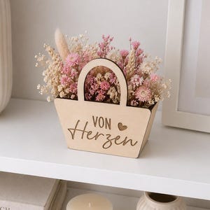 May include: A light-colored wooden basket with a handle, featuring the engraved text "VON Herzen" and a small heart symbol. The basket is filled with a dense arrangement of dried flowers in shades of pink, cream, and beige, including baby's breath.
