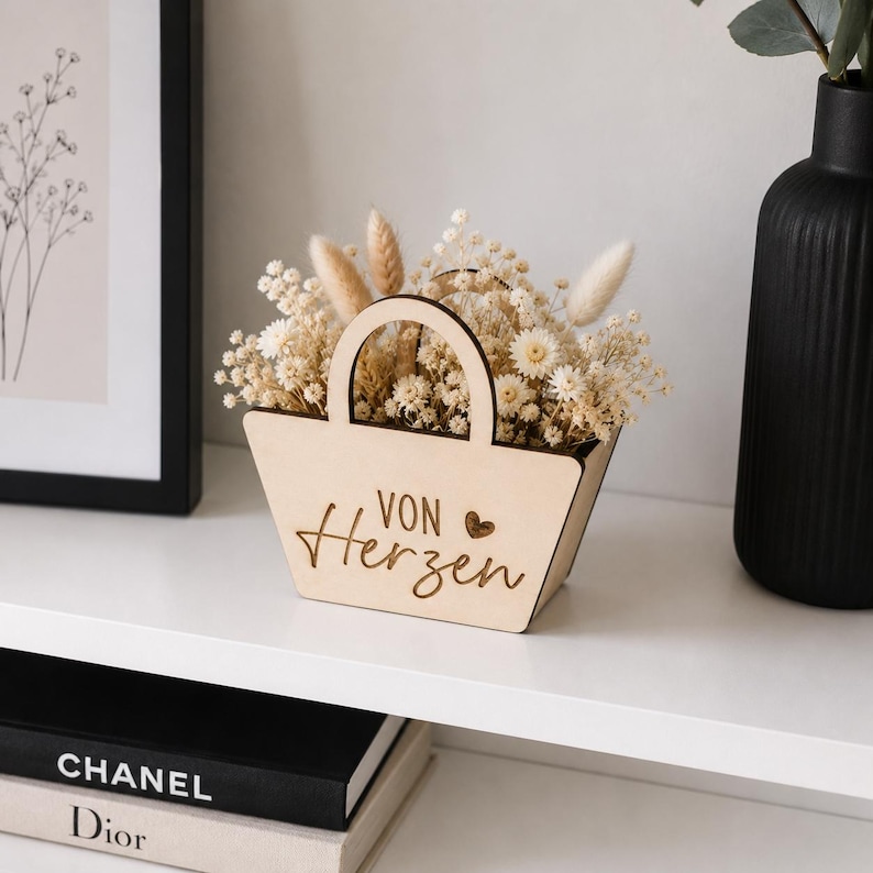 May include: A light-colored wooden basket with a handle, featuring the engraved text "VON Herzen" and a small heart symbol. The basket is filled with an arrangement of dried white flowers, including small blossoms and fluffy, elongated seed heads.