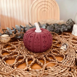 May include: A deep maroon decorative pumpkin featuring a white stem and intricate embossed patterns of hearts and leafy vines carved into its rounded surface. The pumpkin has a textured, ribbed exterior with a matte finish and a symmetrical design.
