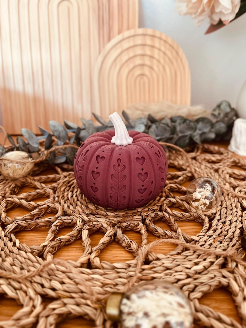 May include: A deep maroon decorative pumpkin featuring a white stem and intricate embossed patterns of hearts and leafy vines carved into its rounded surface. The pumpkin has a textured, ribbed exterior with a matte finish and a symmetrical design.