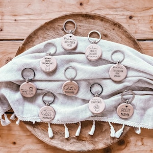 Puede incluir: Una colección de llaveros de madera con aros plateados. Cada llavero tiene un colgante circular de madera con texto grabado y pequeñas ilustraciones. El texto incluye frases como "Mama, wir lieben dich!" y "Beste Mama."