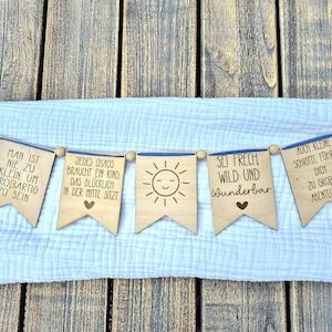 Puede incluir: Una guirnalda de madera con cinco banderines, cada uno con una frase diferente en alemán. Los banderines están ensartados en una cinta azul y tienen un acabado de madera natural. La guirnalda se muestra sobre un fondo de tela blanca.
