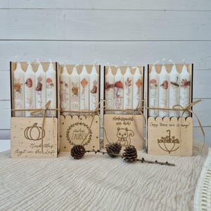 May include: A set of four candle holders with candles, each featuring fall-themed illustrations and text. The wooden holders have decorative cutouts and are tied with twine. The candles display images of pumpkins, umbrellas, and leaves.