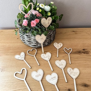 Könnte beinhalten: Eine Sammlung von herzförmigen Holzdekorationen mit Stielen, einige mit Text wie "Mama" und "Beste Mama", auf einer Holzoberfläche angeordnet. Ein Korb mit Blumen mit rosa und weißen Blüten befindet sich im Hintergrund.