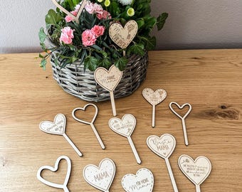 May include: A collection of wooden heart-shaped decorations with stems, some with text like "Mama" and "Best Mom," arranged on a wooden surface. A woven basket of flowers with pink and white blooms is in the background.