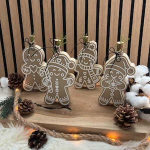 Könnte beinhalten: Vier hellbraune Holzfiguren in Lebkuchenmann-Form mit weiß bemalten Details, jeweils mit einem kleinen Korken und etwas Grün versehen. Die Figuren zeigen verschiedene Designs wie Mützen, Schals und Zuckerstangen, gebunden mit schwarzer Schnur.