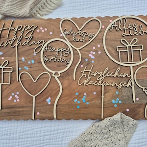 May include: A selection of wooden birthday cake toppers with phrases like "Happy Birthday" and "Alles Liebe". The toppers feature balloon, heart, and gift box designs, arranged on a wooden board. Ideal for a birthday celebration.