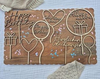 May include: Wooden birthday cake toppers with various designs and phrases, including "Happy Birthday" and "Alles Liebe". The toppers are in the shape of balloons, hearts, and gift boxes, and are set on a wooden board.